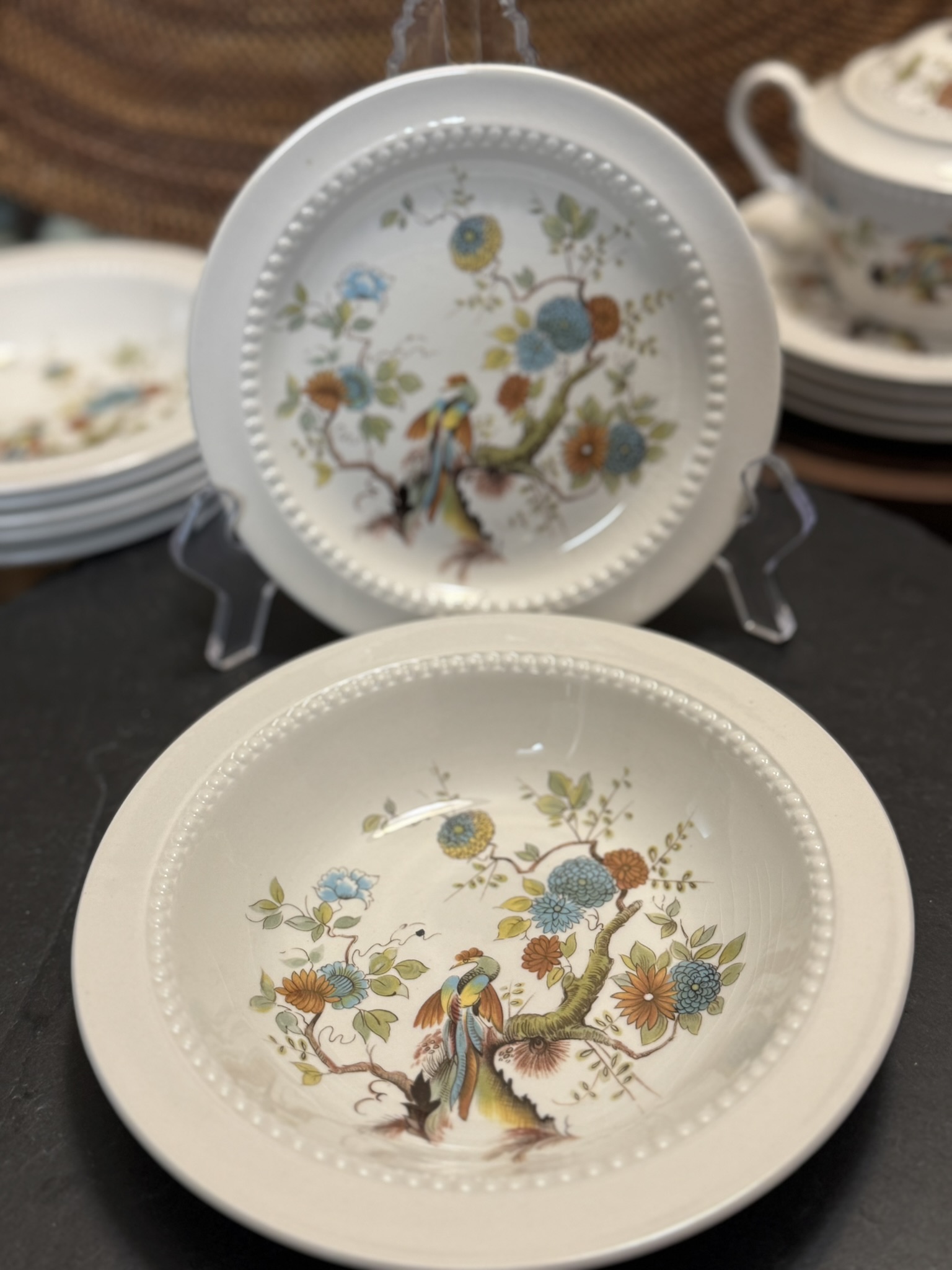 Two white ceramic plates with a beaded rim feature a colorful floral and bird design in orange, blue, yellow, and green. One plate is displayed upright on a stand, while the other lies flat in front. Stacked plates and a teacup are in the background.