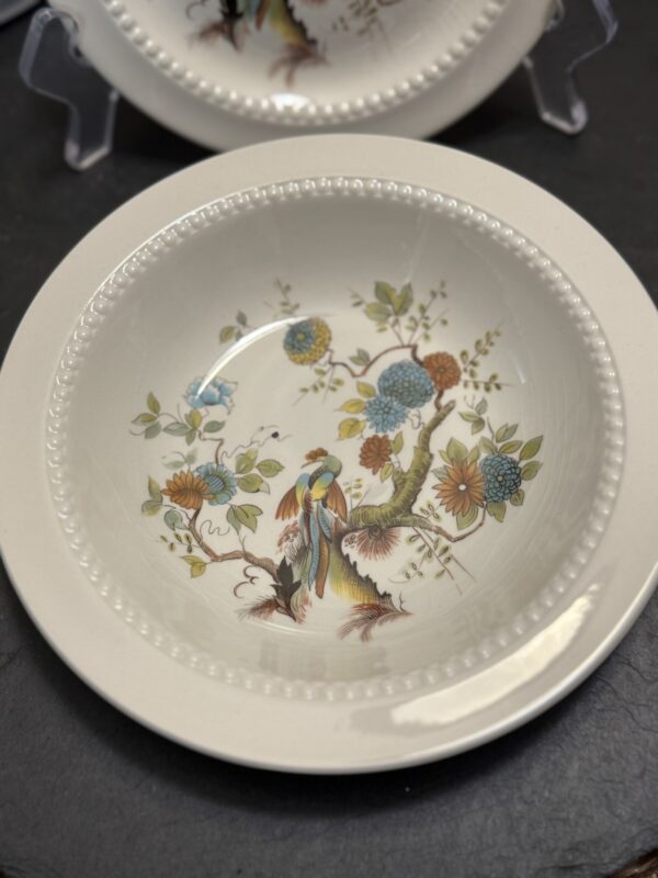 A white ceramic bowl features a raised beaded rim and is decorated with a colorful bird perched on a flowering branch, surrounded by blue, orange, and yellow flowers and green leaves. Another matching plate is partially visible in the background.