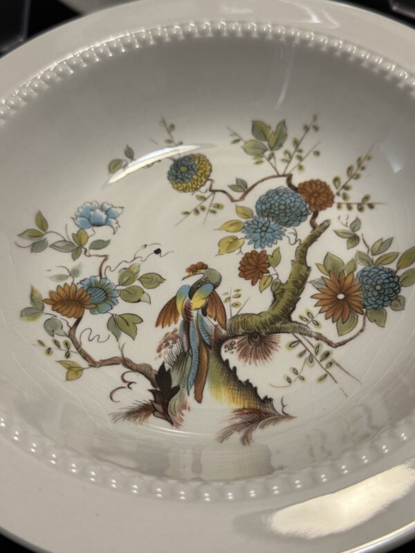 A white ceramic plate with a beaded rim features a detailed, colorful illustration of a bird perched on a tree branch, surrounded by blue, orange, and yellow flowers with green leaves. The design is intricate and has an elegant, vintage style.
