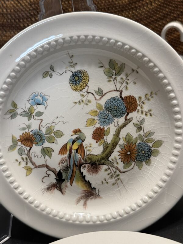 A decorative porcelain plate features a colorful bird perched on a tree branch among large blue, orange, and yellow flowers with green leaves. The plate has a beaded rim and a fine crackle texture, giving it an antique appearance.