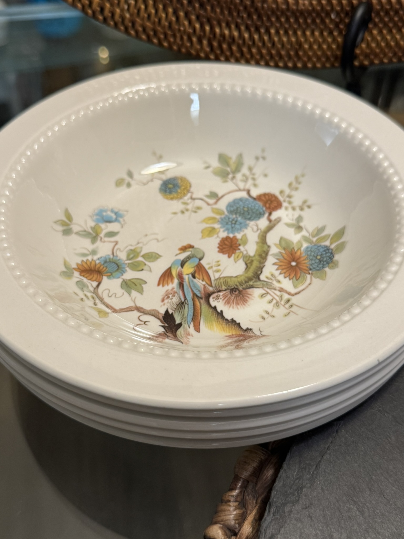 A stack of shallow white bowls, the top bowl featuring a colorful design with a bird perched on a branch, surrounded by blue, orange, and yellow flowers with green leaves. The bowl has a beaded rim detail and sits on a glass surface.