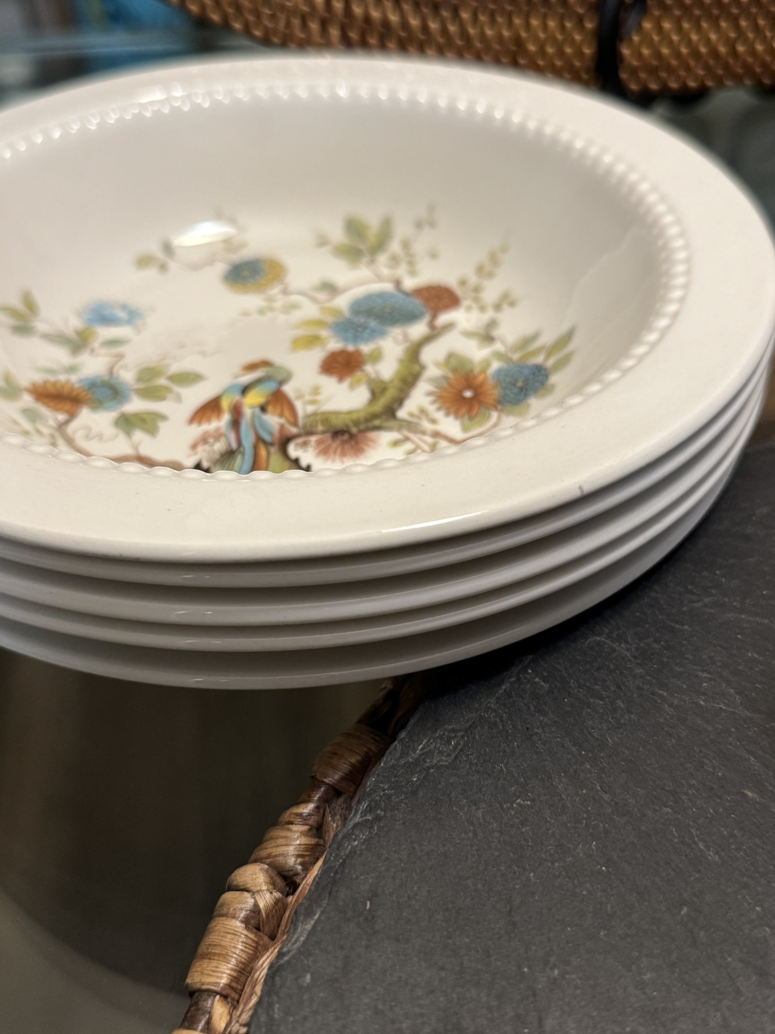 A close-up of a stack of five cream-colored ceramic bowls with a beaded rim and floral pattern inside, featuring blue, red, and green colors. The bowls rest on a black slate surface beside a woven basket edge.