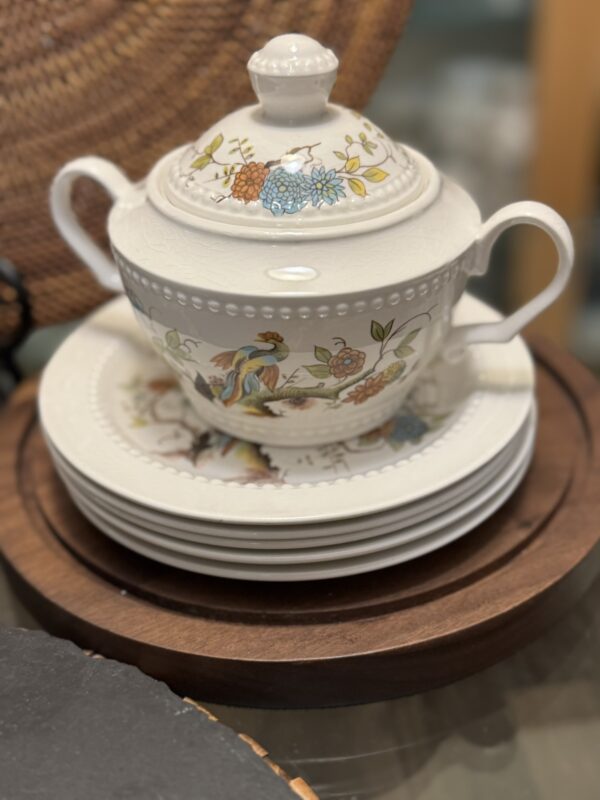 A white ceramic tureen with two handles and a lid, decorated with colorful floral and bird patterns, rests on a stack of matching plates atop a round wooden tray. A woven basket is partially visible in the blurred background.