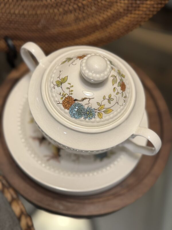 A white ceramic teapot with a lid and handles sits on a matching plate. Both pieces feature a delicate floral pattern in orange, blue, and green. The set rests on a round wooden tray, and a woven surface is partially visible in the background.