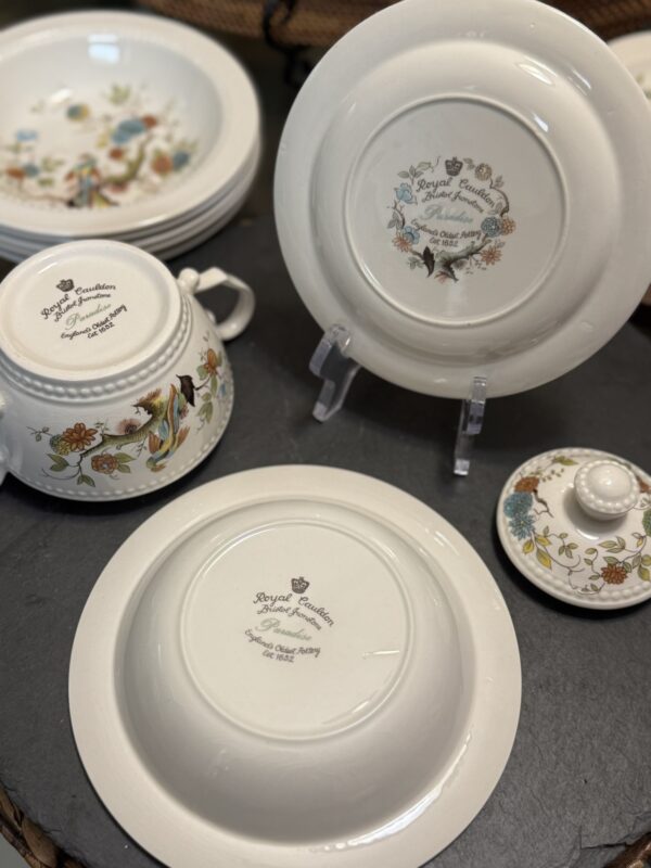 A close-up of Royal Cauldon porcelain dishware displays various pieces with floral and bird designs. The bottoms of a bowl and a plate are visible, showing the brands stamp and motto. Other decorated dishes, including a teapot and lid, are also shown.