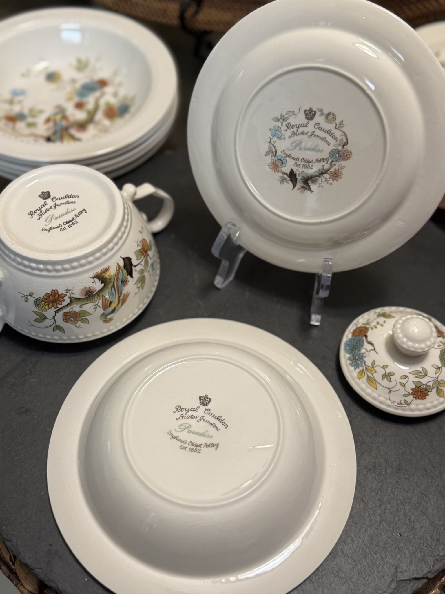 A close-up of Royal Cauldon porcelain dishware displays various pieces with floral and bird designs. The bottoms of a bowl and a plate are visible, showing the brands stamp and motto. Other decorated dishes, including a teapot and lid, are also shown.