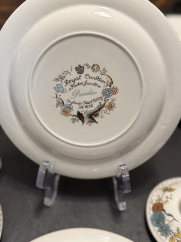 A white porcelain plate on a display stand shows the Royal Cauldon Bristol Ironstone logo with floral illustrations and text including Perception and England’s Oldest Pottery Est. 1652 in elegant script and print. Other plates are partially visible nearby.