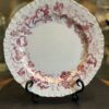 A white porcelain plate with a scalloped, ruffled edge, decorated with a maroon grapevine pattern along the border. The plate rests upright on a black metal stand, and glass shelves with other decorative items are blurred in the background.