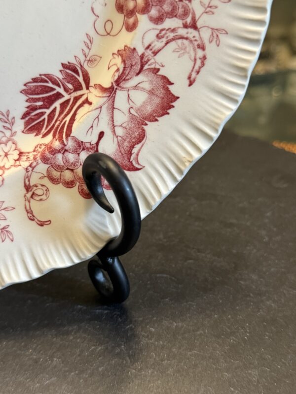 A close-up of a white ceramic plate with red floral and leaf patterns, sitting on a black metal display stand. The plate has a fluted, scalloped edge and rests on a dark, flat surface. Only part of the plate and stand are visible.