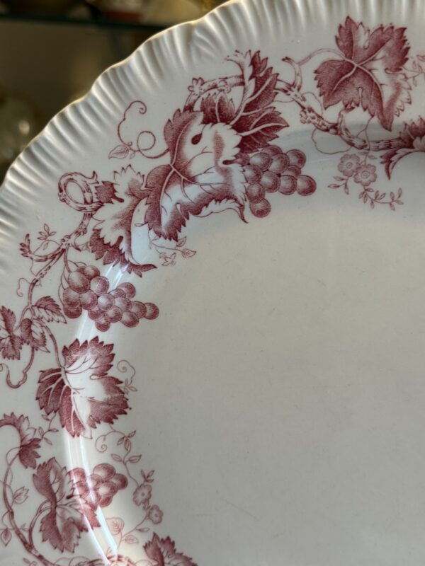 A close-up of a white ceramic plate with a scalloped, ridged edge. The rim features intricate maroon grapevine patterns with clusters of grapes, leaves, and tendrils. The center of the plate is plain and unadorned.