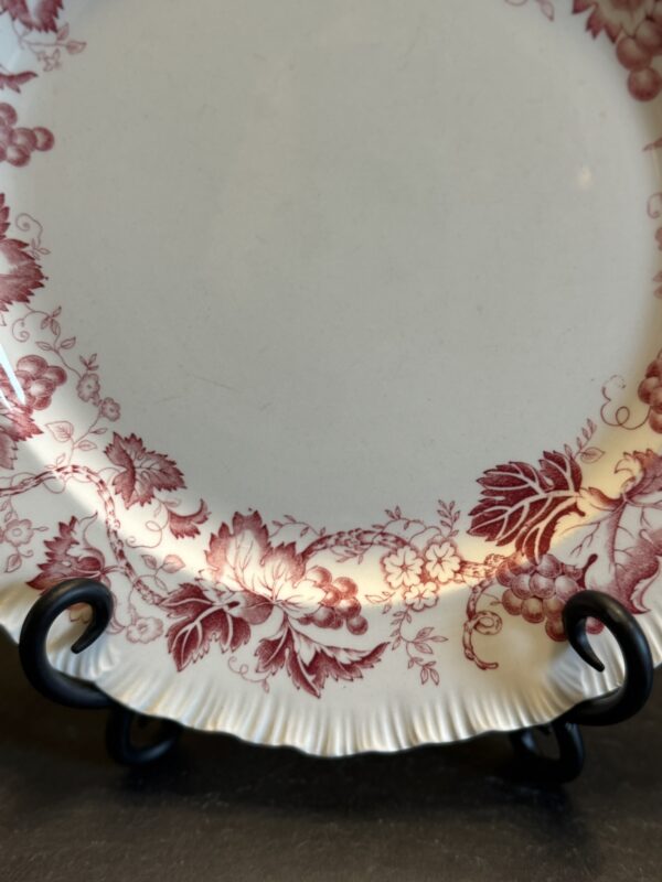 A close-up of a vintage cream-colored plate with a scalloped edge, decorated with intricate red floral and grapevine patterns around the rim. The plate is displayed on a black metal stand. The center of the plate is plain.