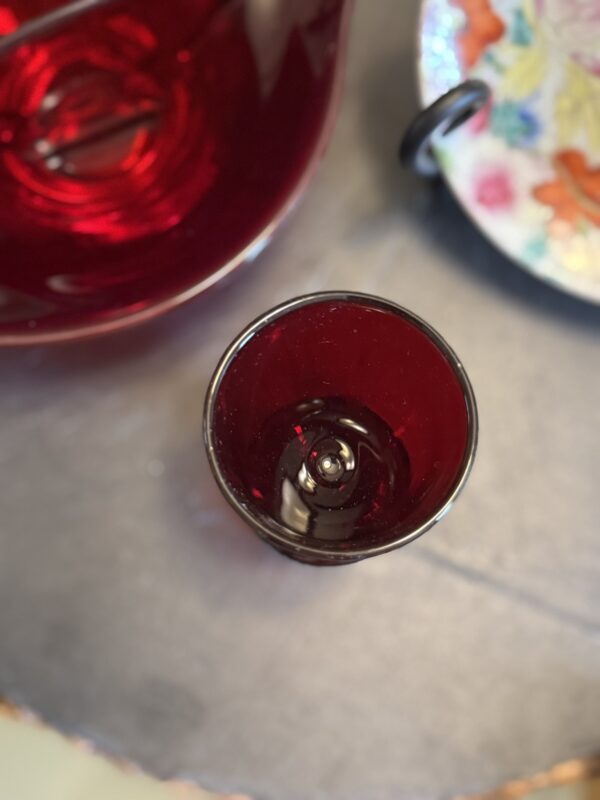 A close-up view of a small, deep red glass cup placed on a gray surface. Part of a matching red glass pitcher is visible to the left, and the edge of a colorful floral-patterned plate appears on the right side of the image.