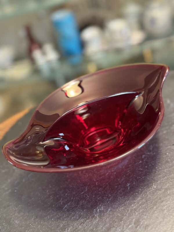 A deep red, glass decorative bowl with a wavy, asymmetric rim sits on a gray textured surface. The background is softly blurred, revealing shelves with various glassware and objects in different colors.