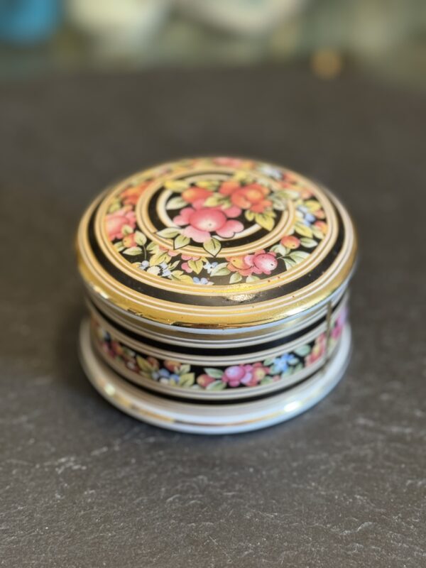 A small, round trinket box with a floral design. The lid and sides are decorated with colorful flowers and green leaves on a black background, accented with gold trim. The box sits on a dark, textured surface.