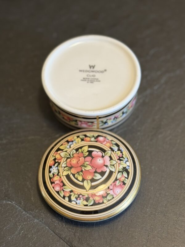 A round Wedgwood ceramic box on a dark surface, with the lid off and set beside it. The lid features a colorful, detailed fruit and floral pattern on a black background, while the inside of the box is plain and marked “WEDGWOOD” and “ENGLAND.”.