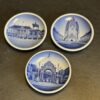 Three small round porcelain plates with blue and white illustrations of historic European buildings and monuments are arranged on a dark, textured surface. Each plate features a different architectural scene in a vintage postcard style.