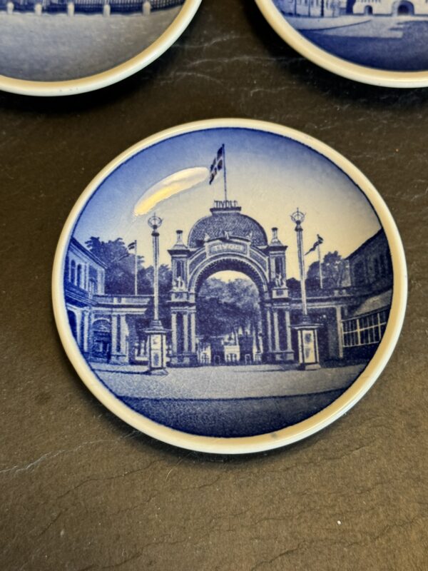 A round ceramic coaster with a blue and white illustration of a grand arched entrance labeled Tivoli, flanked by street lamps and buildings, with a flag flying above the arch and trees visible through the entrance.
