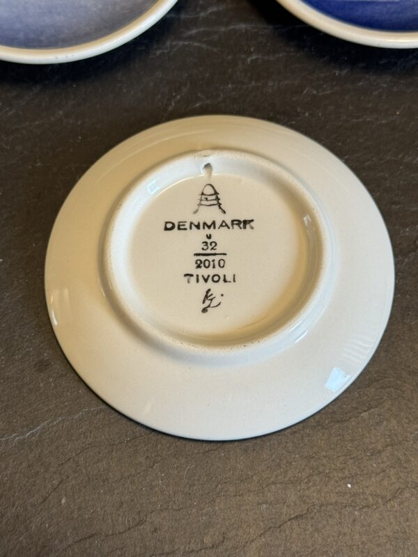 The underside of a ceramic plate shows a maker’s mark: DENMARK, the number 32, year 2010, and TIVOLI. There is a small hanging hole, a logo above the text, and a signature at the bottom. The plate rests on a dark textured surface.