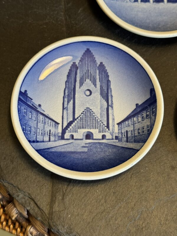 A round ceramic plate features a blue-toned illustration of Grundtvig’s Church, a tall, stepped Gothic-style building flanked by two symmetrical rows of houses, all under a clear sky. The plate has a white rim and sits on a dark surface.