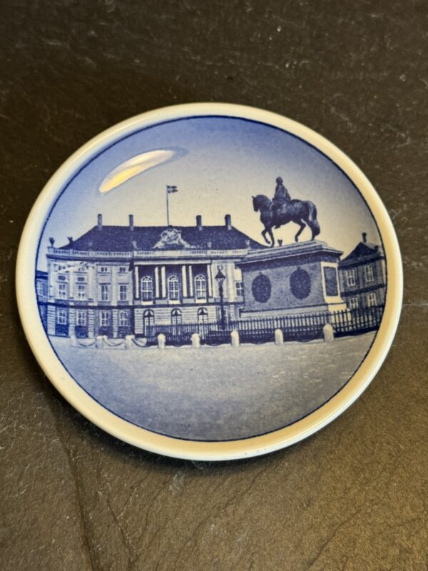 A small round ceramic plate with a blue illustration of a grand building and an equestrian statue in front, featuring classic European architecture and a flag on the roof, set against a dark textured background.