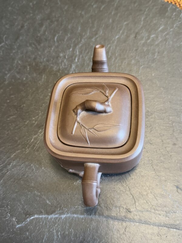 A small, square, brown teapot with a bamboo design on its lid sits on a dark, textured surface. The teapot has a handle and spout perpendicular to each other and a knob on top shaped like a bamboo stalk segment.