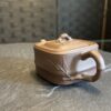 A small, square, brown clay teapot with a bamboo-shaped handle and spout sits on a dark stone surface. The teapot’s lid has a curved knob, and bamboo leaf designs are etched on its sides. The background is softly blurred with a textured wall.