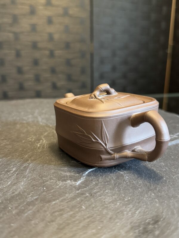 A small, square, brown clay teapot with a bamboo-shaped handle and spout sits on a dark stone surface. The teapot’s lid has a curved knob, and bamboo leaf designs are etched on its sides. The background is softly blurred with a textured wall.