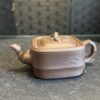 A small, square, brown teapot with a curved spout and handle sits on a dark surface. The lid has a loop handle and bamboo leaf patterns are etched on the lid and body. The background is blurred with a textured, dark wall.