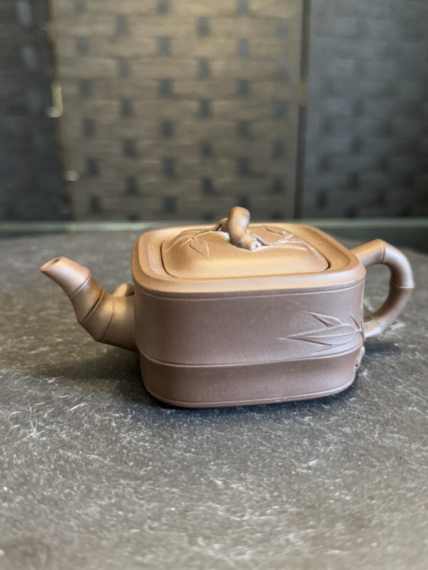 A small, square, brown teapot with a curved spout and handle sits on a dark surface. The lid has a loop handle and bamboo leaf patterns are etched on the lid and body. The background is blurred with a textured, dark wall.