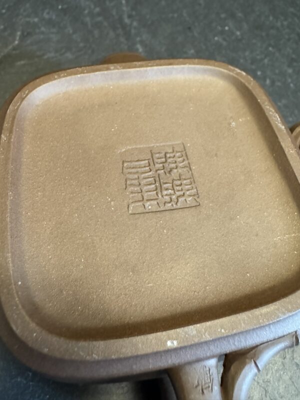 Close-up of the base of a brown clay teapot with a square seal featuring intricate Chinese characters engraved in the center. The teapot surface has a matte texture and subtle color variations, and the background appears to be a dark, smooth surface.