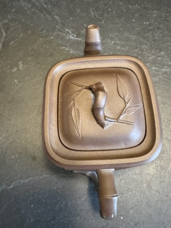 A square, brown clay teapot with a raised bamboo branch design on the lid. The teapot has short spout and handle, both rounded, and sits on a dark stone surface. The lids bamboo motif includes leaves and a textured stalk.