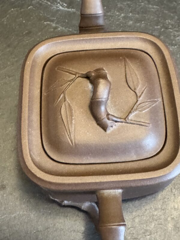 A close-up view of a brown, square teapot lid featuring a raised bamboo branch with two leaves and a bamboo shoot-shaped handle, all crafted in clay. The surface has a matte finish and sits on a dark, textured background.