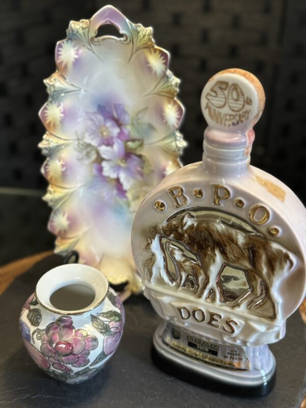 Three vintage ceramic items: a floral-painted plate with gold edges, a pink flowered vase, and a bottle shaped like two elk with “B.P.O. DOES 50th Anniversary” on the stopper. All are displayed on a dark surface with a blurred dark background.
