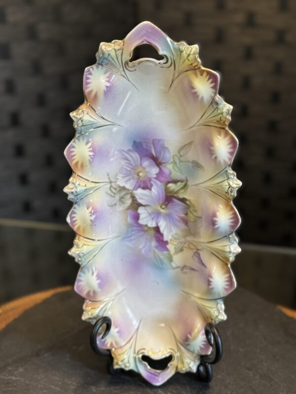 An ornate, pastel-colored porcelain tray with scalloped, floral-edged sides and two cut-out handles. The surface features hand-painted purple flowers and green leaves, with soft gradients of pink, yellow, and light blue, displayed upright on a black stand.