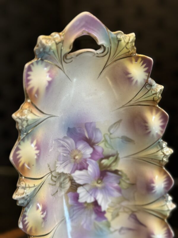 An ornate, leaf-shaped porcelain plate features embossed edges with a gradient of pastel yellows, purples, and greens. The center displays painted purple and white flowers with green leaves. The top of the plate has a cutout handle. Background is blurred.