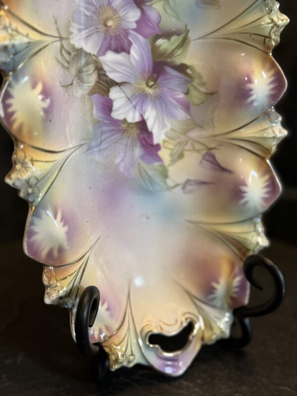 An ornate, scalloped porcelain plate with pastel hues of lavender, yellow, and green. The upper section is decorated with detailed purple and white flowers and green leaves. The plate is displayed on a metal stand against a dark background.
