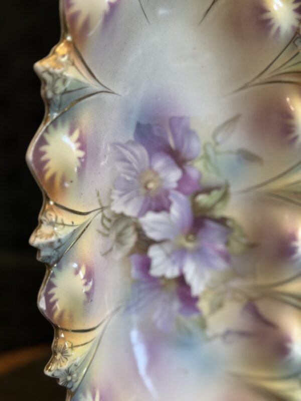 A close-up of a porcelain vase with a scalloped, ruffled edge. The vase features a pastel gradient of purple, pink, and cream tones, detailed with hand-painted purple and white flowers and green leaves, accented by gold outlines and delicate texture.