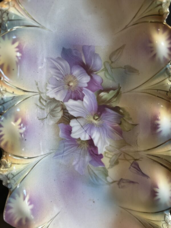 A decorative porcelain plate features hand-painted purple and white flowers with green leaves. The plate has a scalloped, ornate rim with pastel accents of pink, blue, and gold around the edges, framing the central floral design.