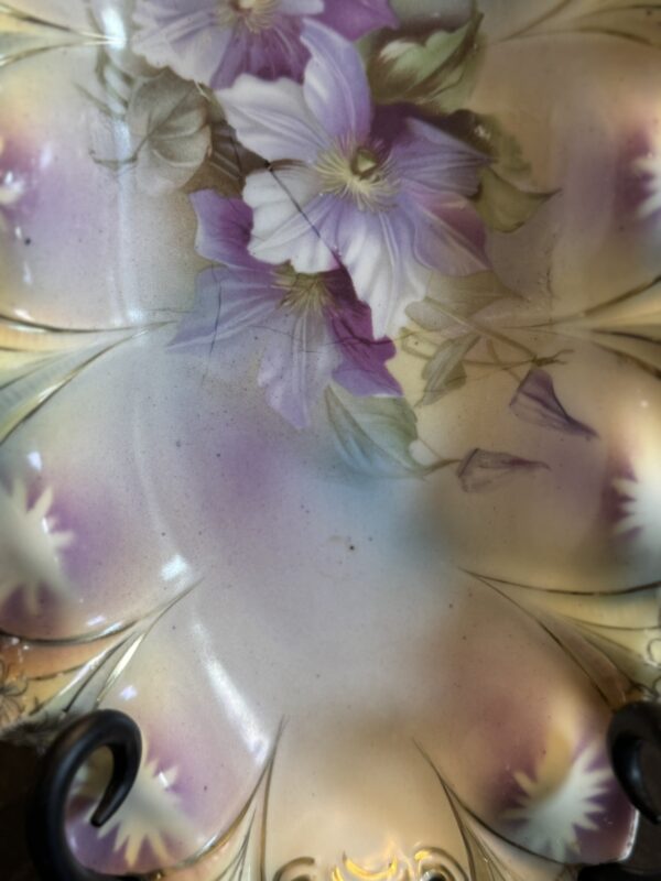 An ornate porcelain plate with scalloped edges, decorated with hand-painted purple and white flowers and green leaves. The plate features soft pastel shades and intricate gold accents along the rim, creating an elegant and vintage appearance.