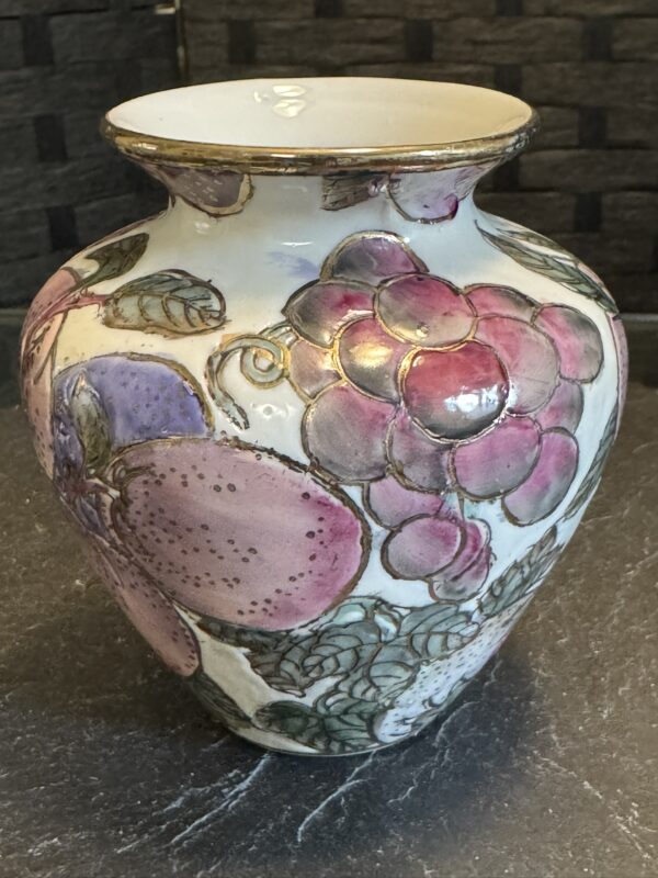 A ceramic vase with a glossy finish, featuring hand-painted fruit motifs including purple grapes and plums, green leaves, and delicate detailing. The vase sits on a dark stone surface with a textured, dark brick background.
