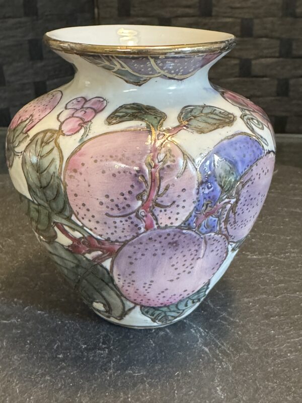 A ceramic vase with a wide mouth and rounded body, decorated with hand-painted pink and purple peaches, green leaves, and gold accents. The background is a white glaze, and the vase sits on a gray stone surface with a textured wall behind it.