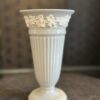 A tall, white ceramic vase with vertical ridges and a flared rim is decorated with a raised floral vine pattern near the top. The vase stands on a round base and is photographed against a blurred, dark background on a textured surface.