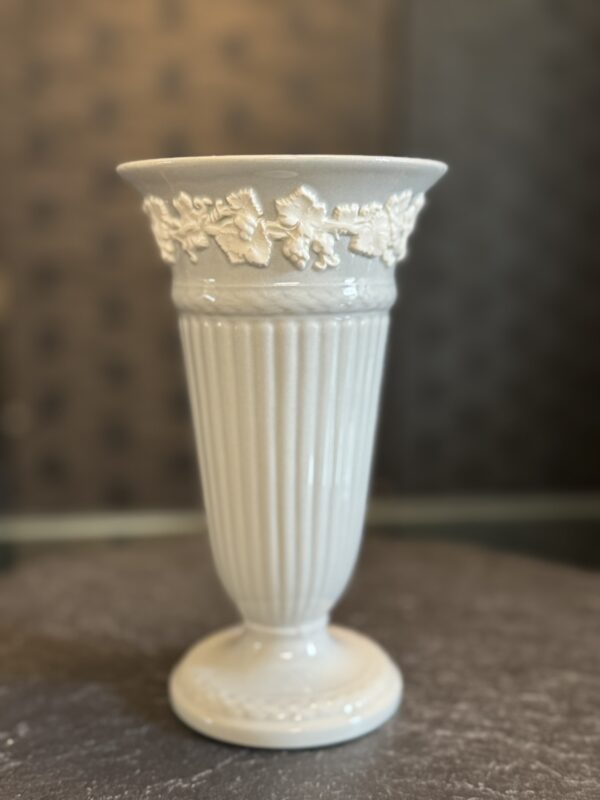 A tall, white ceramic vase with vertical ridges and a flared rim is decorated with a raised floral vine pattern near the top. The vase stands on a round base and is photographed against a blurred, dark background on a textured surface.