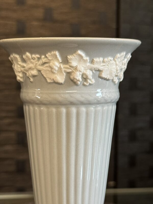 A close-up of a white ceramic vase featuring a fluted body and a decorative band of raised grapevine motifs, including leaves and grape clusters, near its flared rim. The background is blurred and dark, highlighting the vase’s details.
