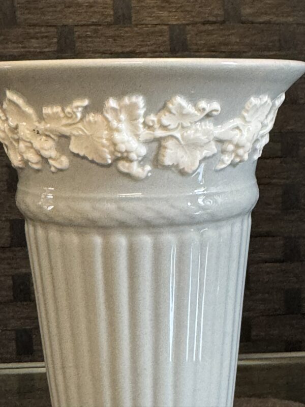 A close-up of a tall, glossy, light gray ceramic vase with vertical ribs and an ornate band of raised vine and leaf decorations near the flared rim. The background is blurred, showing a textured brown wall.