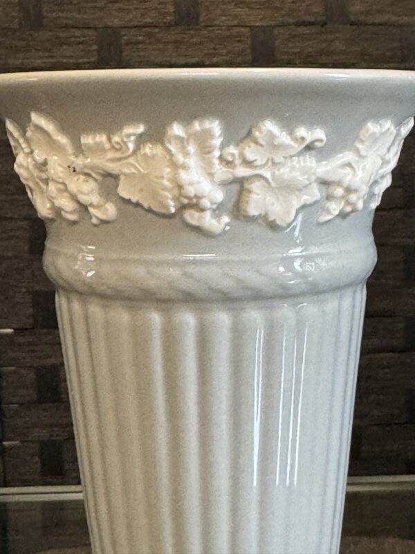 A close-up of a white ceramic vase with a glossy finish. The top edge features raised, decorative grapevine and leaf motifs, while the body below has vertical fluted ridges. The background is slightly blurred and shows a brown, textured surface.