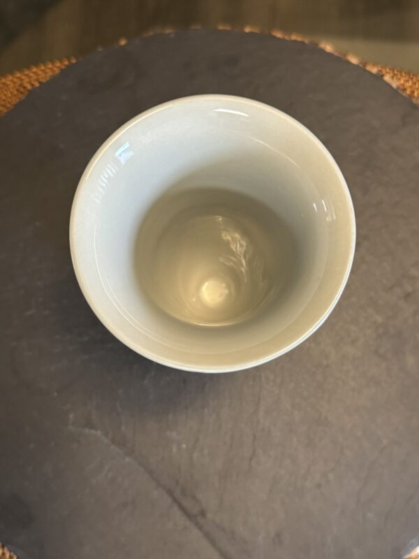 A top-down view of an empty, light gray ceramic cup sitting on a dark, round surface with a textured, brown edge partially visible in the background. The cup has a smooth and glossy finish, reflecting light.