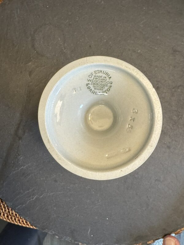 A light gray ceramic bowl sits upside down on a dark surface. The base reveals embossed text: “OF ETRURIA MADE IN ENGLAND WEDGWOOD & CO GARLAND 3203.” The number 315 is also visible on the outer base. Part of a woven placemat is visible in the corner.