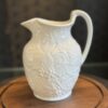 A white ceramic pitcher with a curved handle and embossed grapevine and leaf patterns sits on a round wooden tray. The background is softly blurred, highlighting the intricate detail on the pitcher’s surface.
