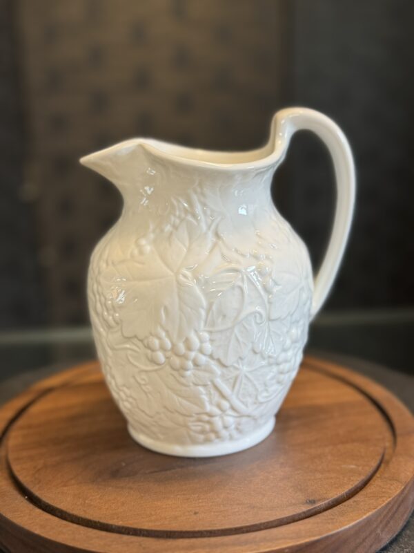 A white ceramic pitcher with a curved handle and embossed grapevine and leaf patterns sits on a round wooden tray. The background is softly blurred, highlighting the intricate detail on the pitcher’s surface.